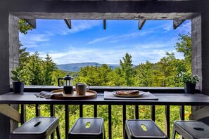 Un balcon avec une table dressée pour le petit-déjeuner, une machine à café et une assiette de nourriture, donnant sur un paysage boisé avec des montagnes à l’horizon.