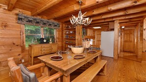 Interior de una cabaña de madera con una mesa de comedor, bancos, una araña y una zona de cocina.