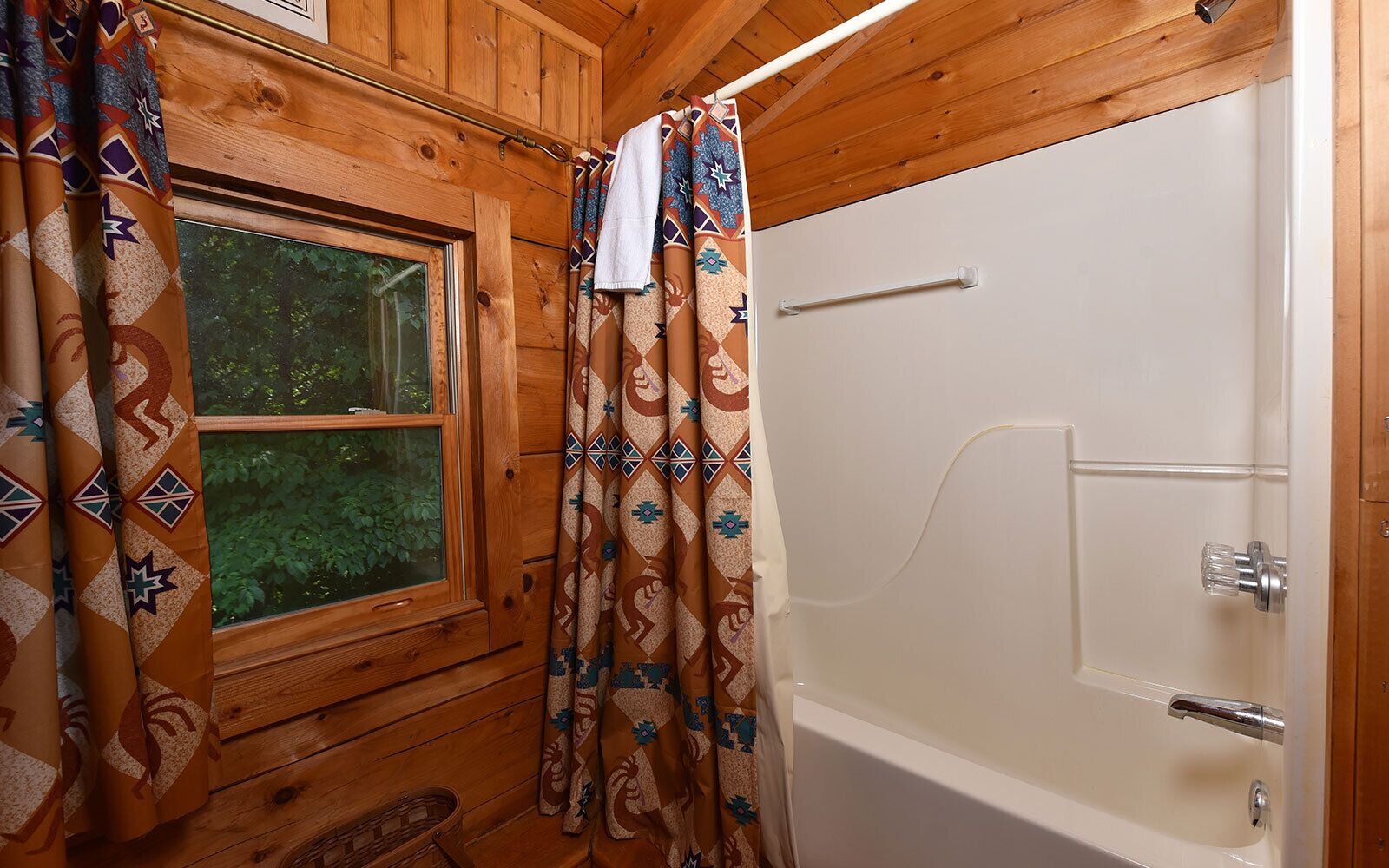 A wooden cabin bathroom with a window, patterned curtains, and a bathtub.