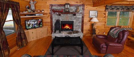 A cozy log cabin interior with a stone fireplace, leather sofa, and a television on a wooden cabinet.