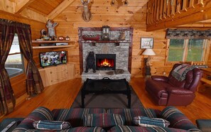 A cozy log cabin interior with a stone fireplace, leather sofa, and a television on a wooden cabinet.