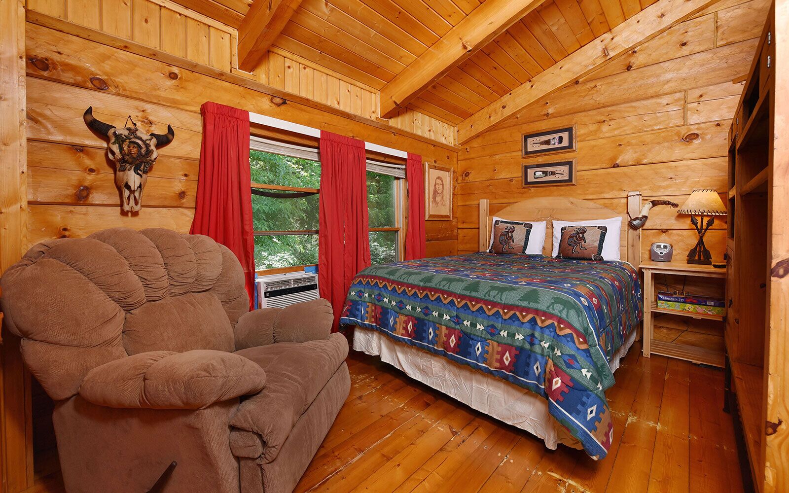 A cozy log cabin bedroom with a large bed, a chair, and a window with red curtains.