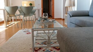 Un salon moderne avec une table basse en verre, un canapé gris et des fauteuils assortis. On aperçoit un meuble blanc et une porte en bois à l’arrière-plan.