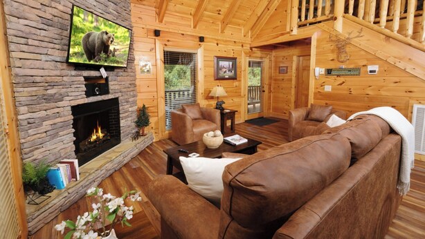 A log cabin interior with a stone fireplace, leather sofas, a television, and a deer head decoration. - Ever After - Community Pool, Spa Tub, Pool Table, Hot Tub, & Smart TV! (Sevierville)