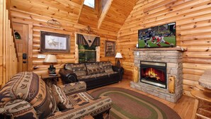A log cabin interior with a fireplace, a television mounted above it, a sofa with patterned cushions, and a framed picture of deer on the wall. - Think a Little Less - Air Hockey, Gas Fireplace (Sevierville)