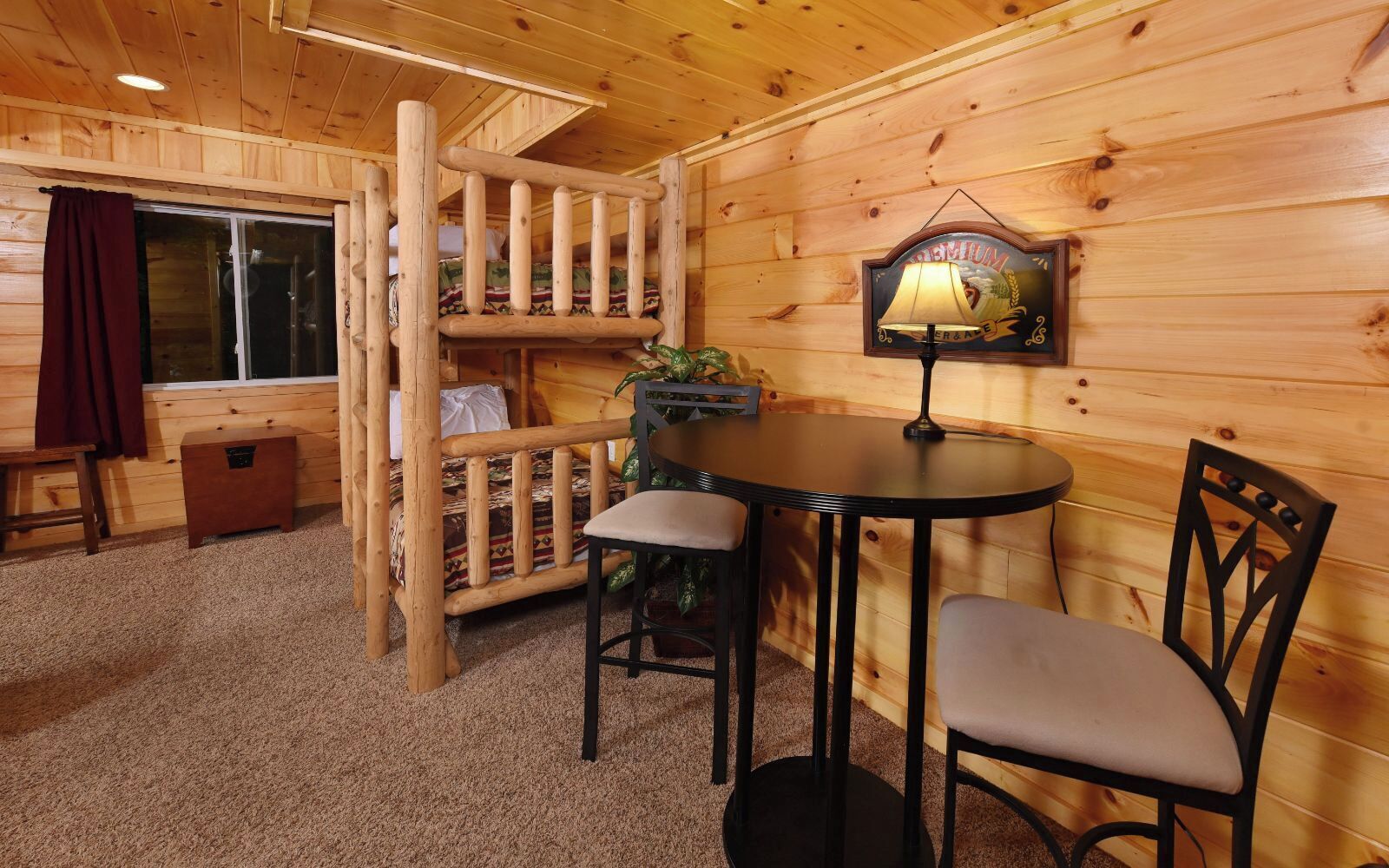 A cozy cabin interior with a bunk bed, a small table with two chairs, a lamp, and a window with curtains.
