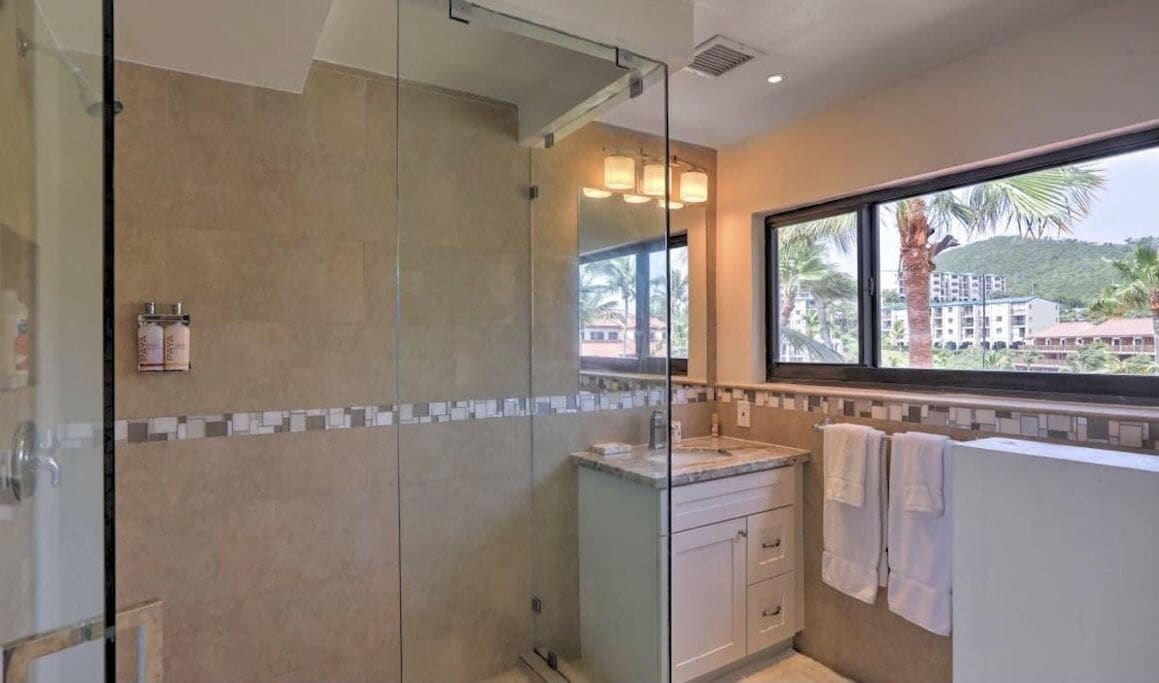 Une salle de bain moderne dotée d’une douche à parois de verre, d’un meuble-lavabo et d’une fenêtre donnant sur des palmiers et des immeubles.