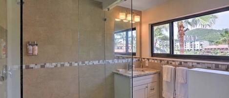 Une salle de bain moderne dotée d’une douche à parois de verre, d’un meuble-lavabo et d’une fenêtre donnant sur des palmiers et des immeubles.