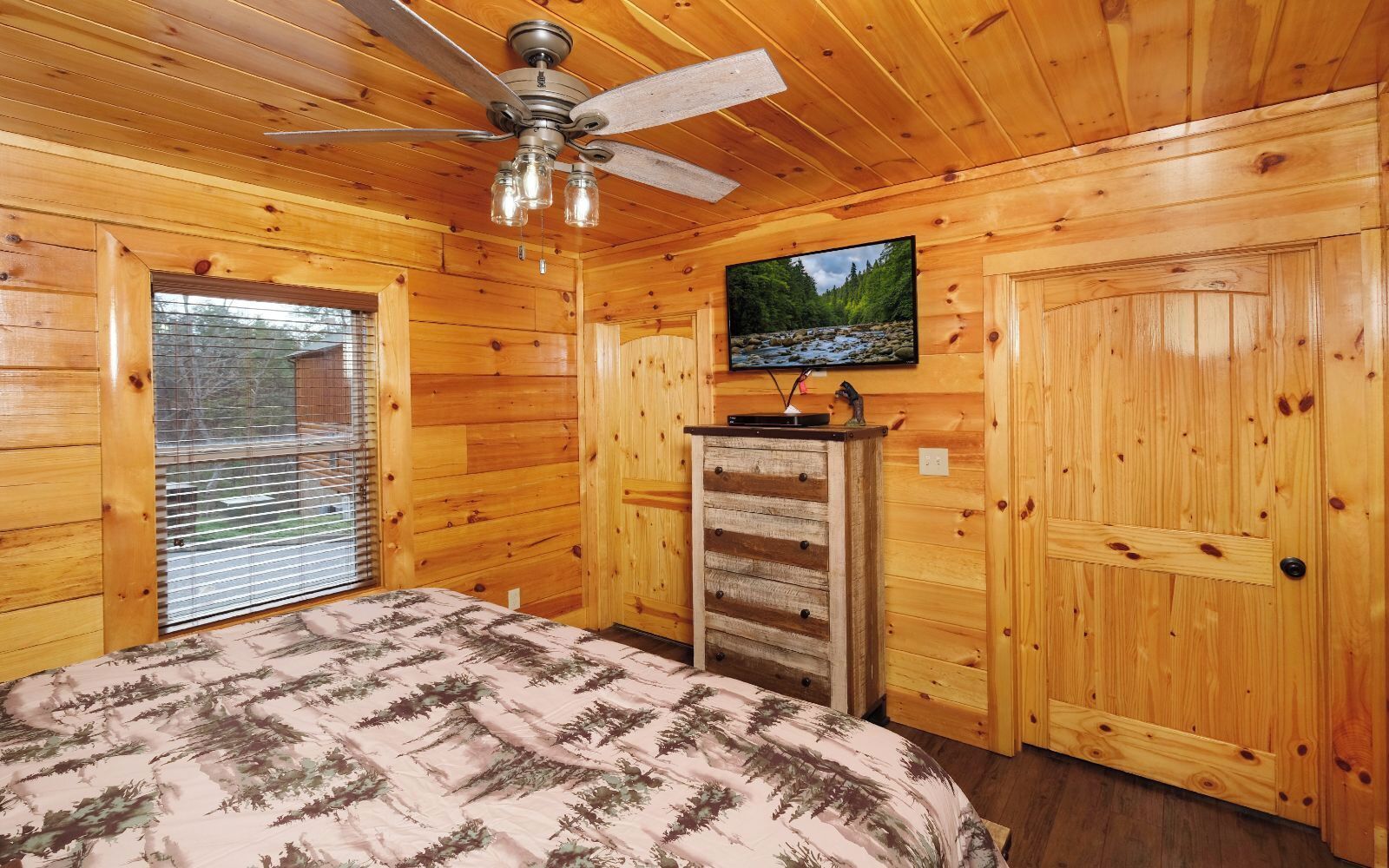 A wooden cabin room with a bed, a ceiling fan, a TV on the wall, and a window with blinds.