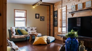 Ein gemütliches Wohnzimmer mit Holzwänden, einer Couch mit gemusterten Kissen, einem Fernseher und einer blauen Vase mit Grünpflanzen.