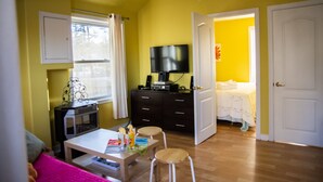 Habitación pequeña con televisión, una mesita de comedor, dos taburetes, una cama y una puerta que da a otra habitación.