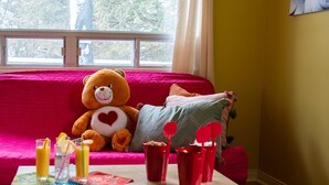 Una habitación acogedora con un ventanal grande, un osito de peluche en un sofá rojo, y una mesa con bebidas y un libro.