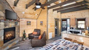 L’intérieur d’une cabane en rondins, avec une cheminée en pierre, un ventilateur de plafond, un téléviseur, un petit sapin de Noël, un canapé et un coin cuisine.
