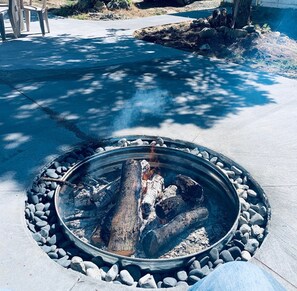 A fire pit with logs and rocks, surrounded by a concrete area with chairs and a tree in the background. - Charming retreat on 20 acres nestled in the woods 1 mile from Lexington  (Croswell)