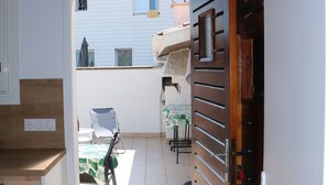 Un couloir étroit mène à un balcon où se trouvent une chaise et une table. Une porte en bois et un appareil électroménager blanc se trouvent sur le côté gauche.