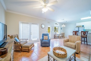 A spacious living room with a television, comfortable seating, a ceiling fan, and a kitchen area in the background.