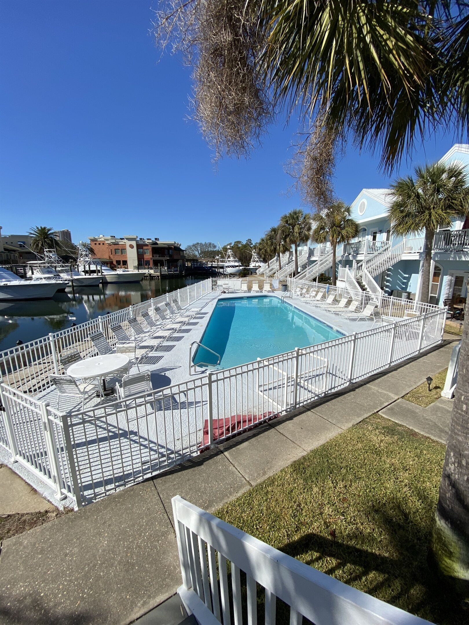 A pool area with a fenced deck, lounge chairs, and a view of boats and buildings in the background.