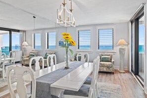 A dining area with a long table, white chairs, and a vase with sunflowers. There are windows with blinds, a chandelier, and a lamp.