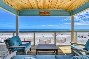 Een overdekt terras met houten plafond, een bord met de tekst 'Welkom op ons terras' en uitzicht op het strand met mensen en parasols.