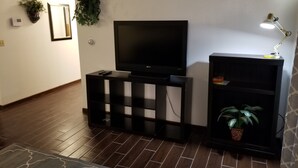 Un salon avec un téléviseur à écran plat posé sur un meuble noir, une petite table avec une lampe et une plante en pot.