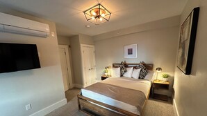 A bedroom with a bed, bedside tables, a flat-screen TV, a ceiling light fixture, and a wall-mounted air conditioner.