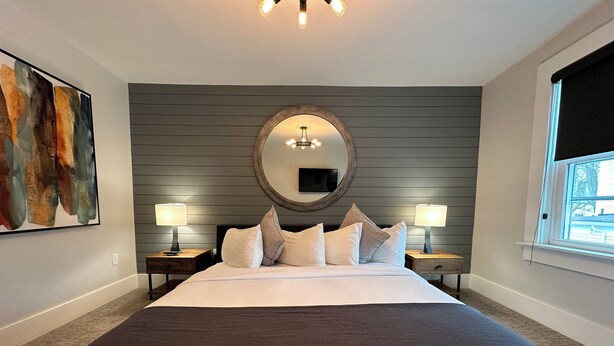 Une chambre à coucher bien aménagée, avec un grand lit, deux tables de chevet avec des lampes, un grand miroir rond et une grande peinture abstraite accrochée au mur.