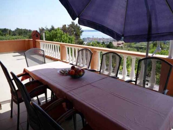 Un balcon avec une table et des chaises, un saladier rempli de fruits, et une vue sur la verdure et un paysage lointain.