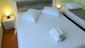 Cama bem arrumada com lençóis e almofadas brancos, cabeceira de madeira e mesa de cabeceira com abajur.