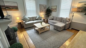 Una sala de estar moderna con dos sofás, una mesa de centro y una lámpara de pie. Hay dos ventanas con persianas, un cuadro en la pared y una planta pequeña sobre la mesa de centro.