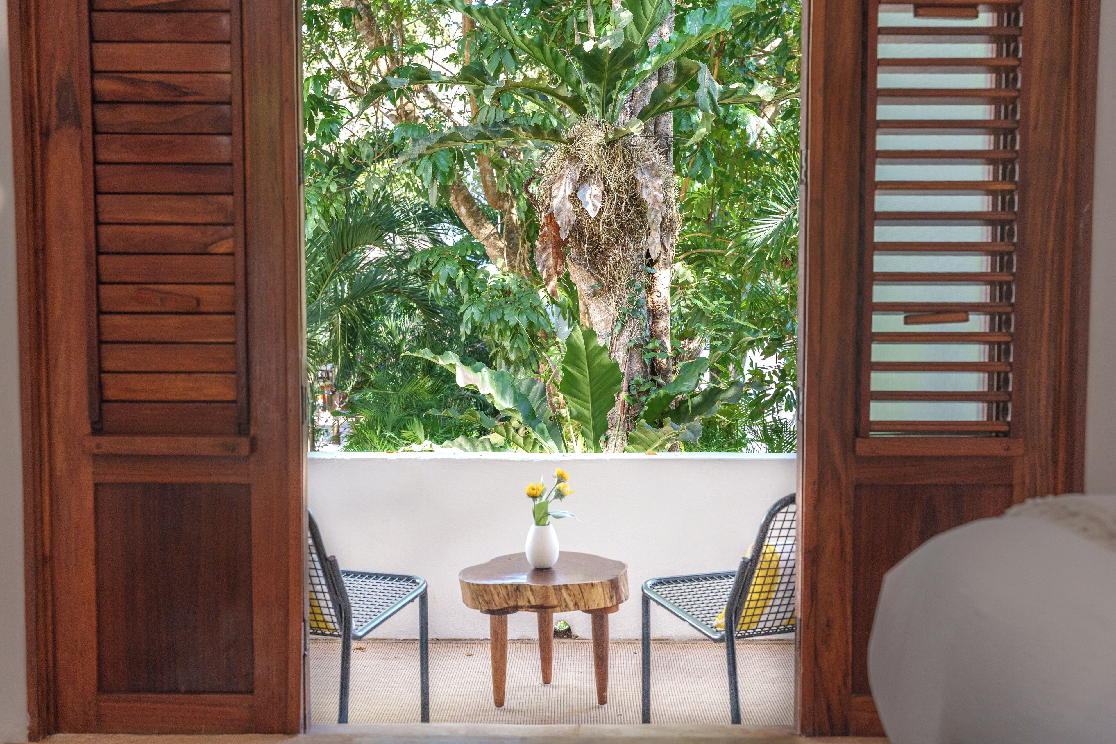 A wooden-framed window with a view of lush greenery and a small round table with a vase of yellow flowers.