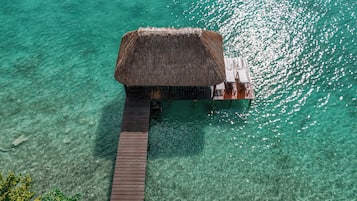 An overwater bungalow with a thatched roof, wooden deck, and a small lounge area with seating.