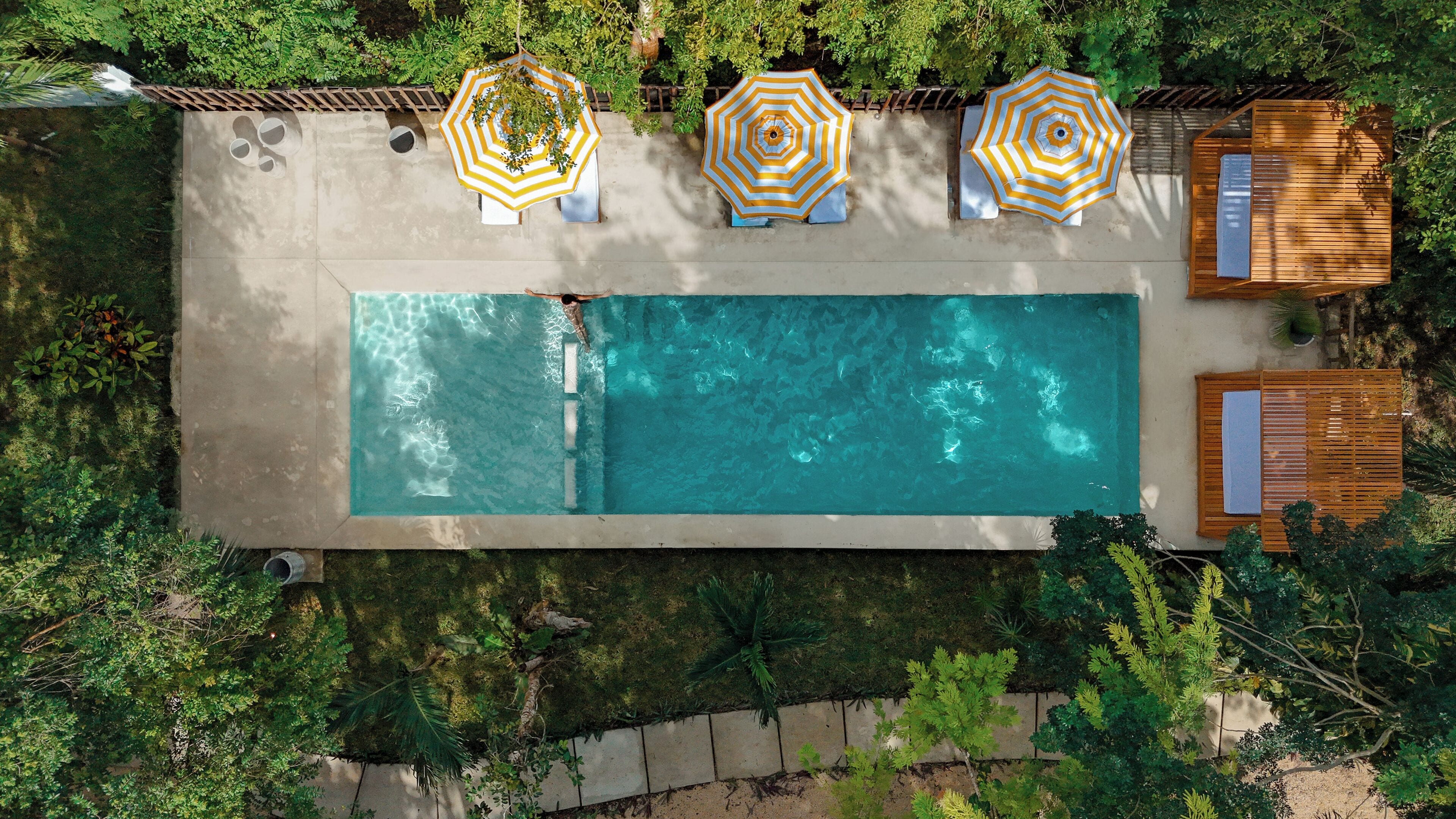 Vista aérea de uma piscina retangular com três guarda-sóis, rodeada por vegetação exuberante e mobiliário de deck em madeira.