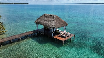 Een houten bungalow boven het water met rieten dak, helderblauw water en een houten terras.