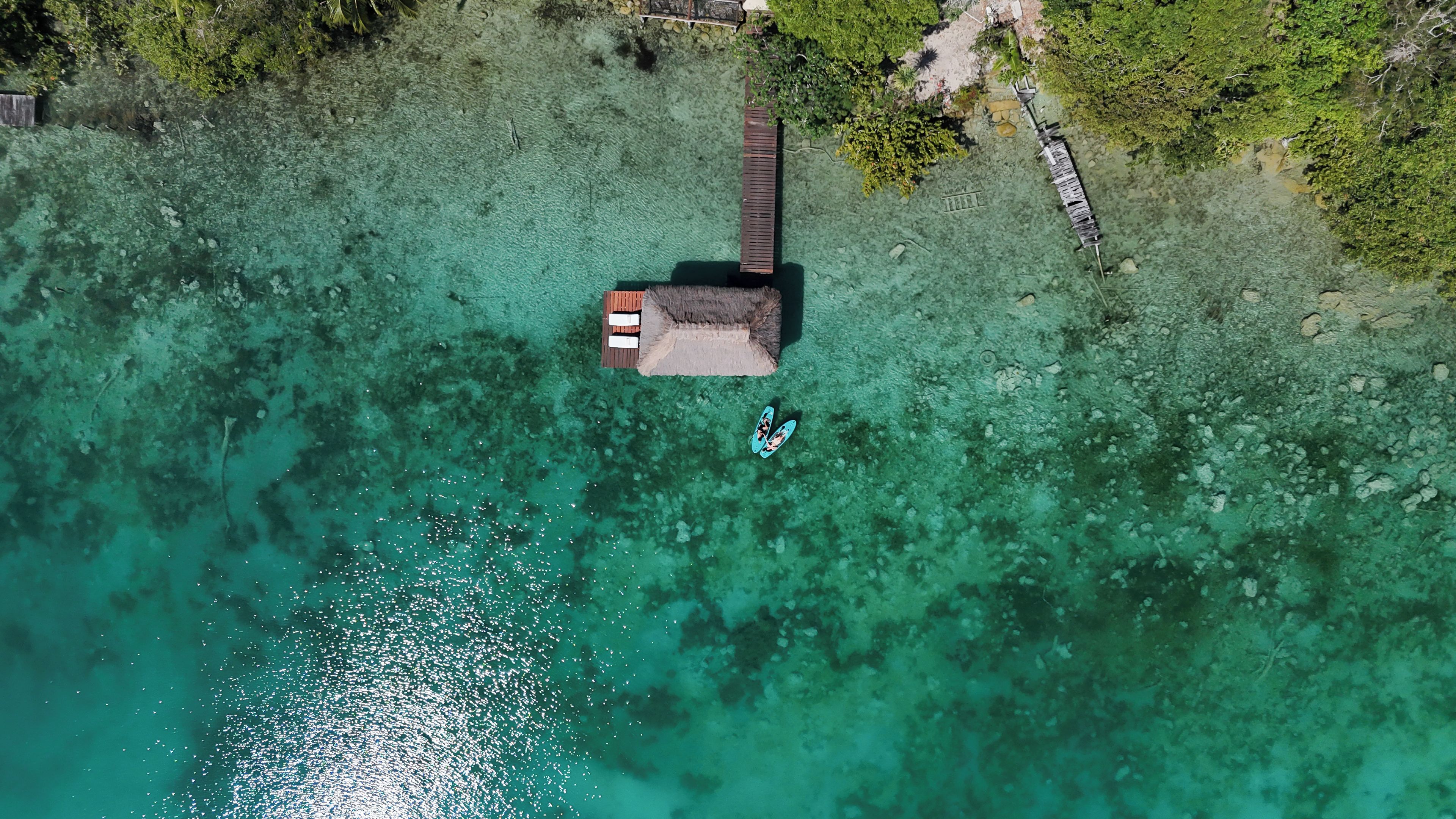An aerial view of a clear turquoise lagoon with a wooden dock leading to a thatched-roof hut.