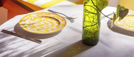 Une table recouverte d’une nappe blanche, une bouteille en verre verte contenant une plante et une assiette ornée d’un motif jaune.
