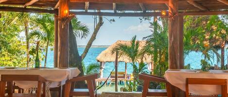 Un área de comedor exterior de madera con mesas y sillas, con vista a una playa con sombrillas de paja y palmeras.