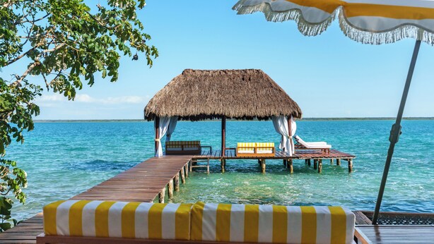 A wooden deck with a thatched-roof hut, wooden lounge chairs, and a small table. - Mikasa Bacalar Hotel Boutique, Lagoon Front (Bacalar)