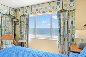 Una habitación con vista al mar, una cama con ropa de cama azul, una silla de mimbre y cortinas con estampado de lunares.