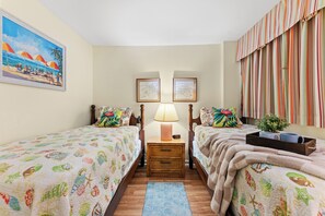 Una habitación con dos camas, una mesita de noche con lámpara, una pequeña mesa y un ventanal con cortinas a rayas.
