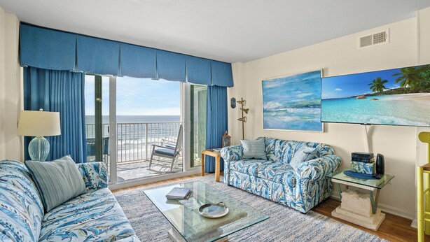 A room with a large window, a sofa, a coffee table, a TV, and a painting of a beach scene.