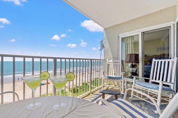 Un balcón con vista a la playa, dos bebidas con rodajas de lima, sillas blancas y una mesa.