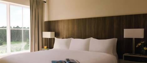 Una habitación de hotel con una cama grande, dos lámparas de noche, una mesita de noche y un ventanal con cortinas.