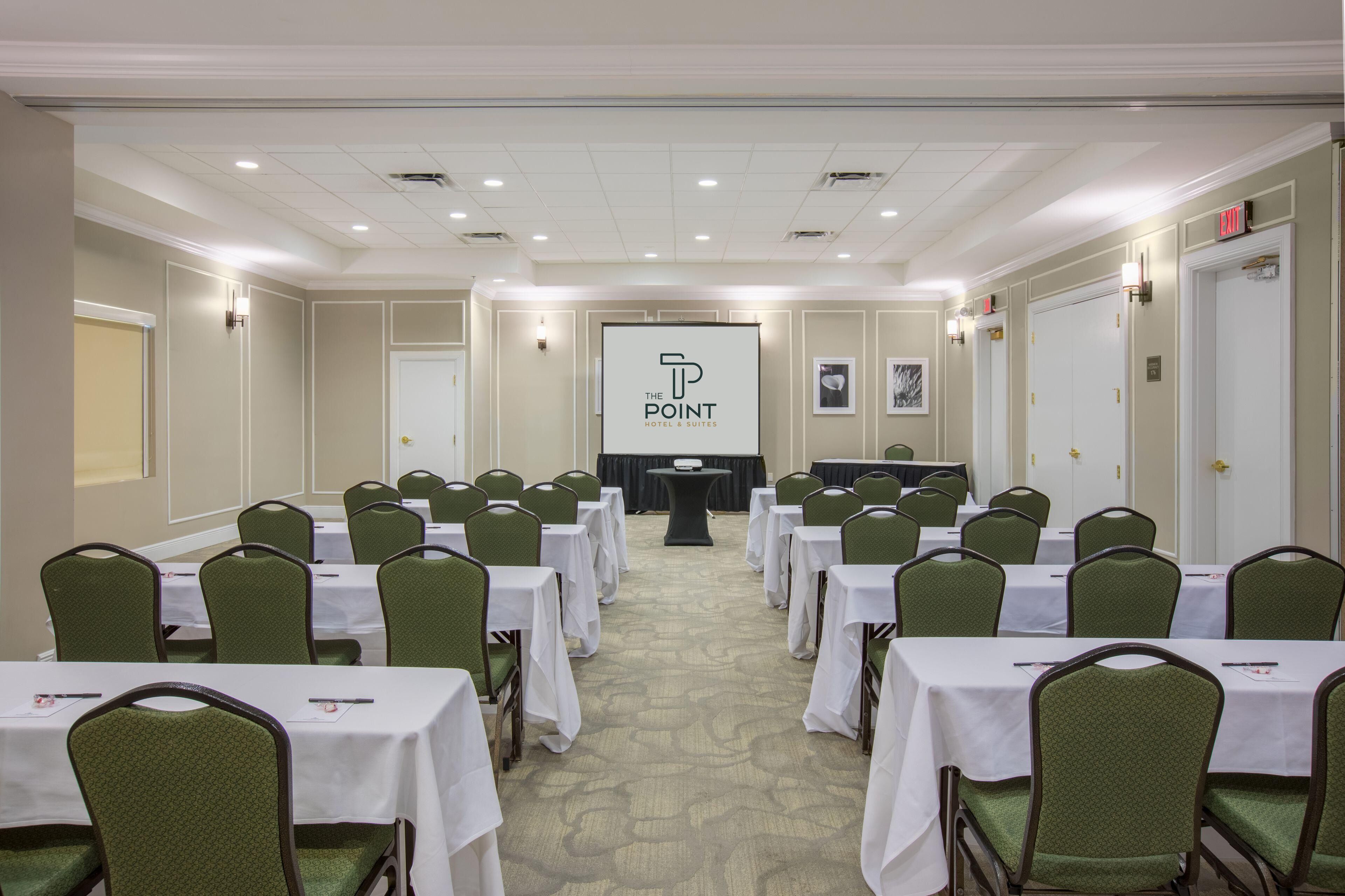 Een congreszaal met rijen tafels en stoelen, een spreekgestoelte en een groot scherm waarop het logo van 'The Point Hotel & Suites' staat.