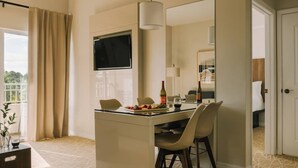 A hotel room with a dining area featuring a glass table, chairs, and a bottle of wine. A large mirror hangs above the table, and a TV is mounted on the wall.