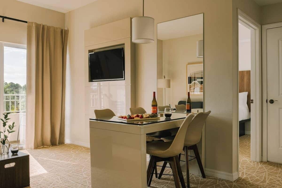 Habitación de hotel con zona de comedor que incluye una mesa de vidrio, sillas y una botella de vino. Un espejo grande cuelga sobre la mesa, y un televisor está montado en la pared.