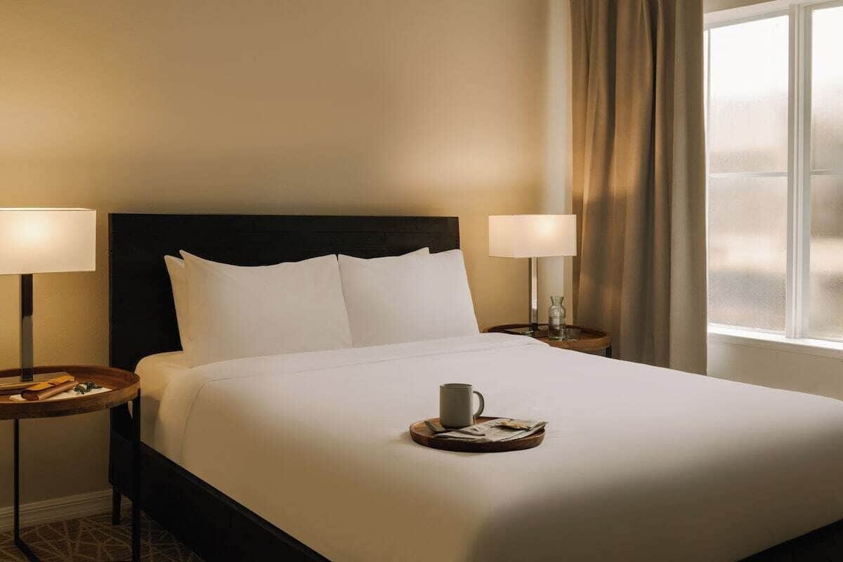 Una cama bien hecha con sábanas blancas, dos lámparas de noche, una mesita de noche con una taza y un libro, y una ventana con cortinas.