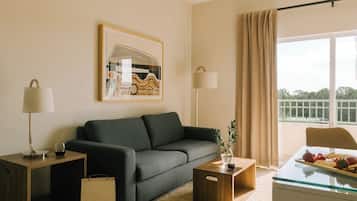 Un salon moderne avec un canapé vert foncé, une table basse en verre et une table d’appoint avec une lampe. Un tableau encadré est accroché au mur, et il y a une fenêtre avec des rideaux.