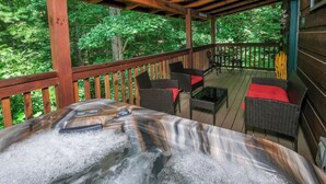 A wooden deck with a hot tub, wicker furniture, and a view of a forest.