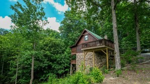 A cabin with a stone foundation and a wooden deck, surrounded by dense forest.