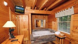 A wooden cabin bathroom with a stone bathtub, wooden walls, a television mounted above the door, and a window with blinds.